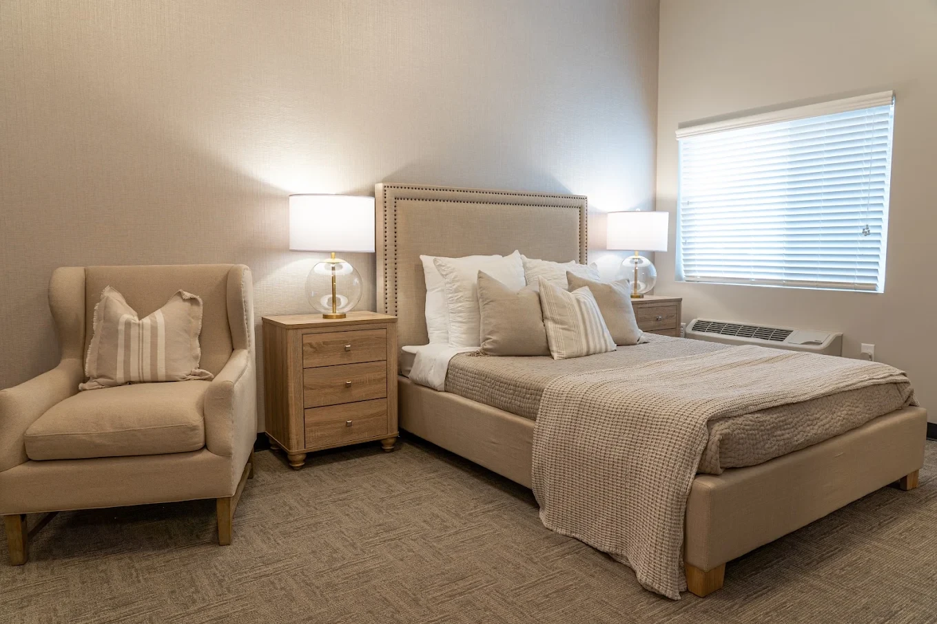 A furnished resident apartment with a queen bed, armchair, and large window