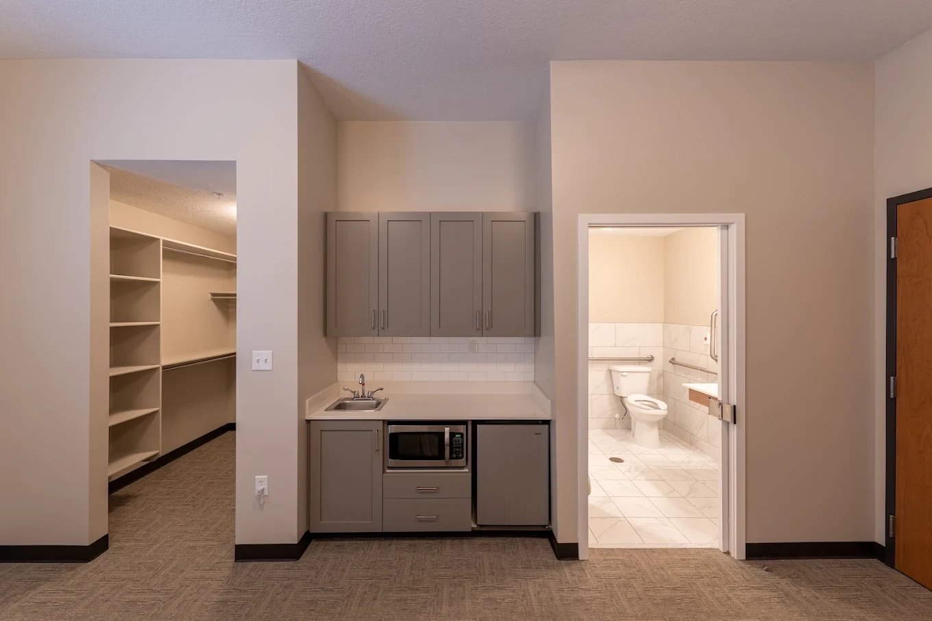 An apartment kitchenette with sink, microwave, and mini fridge, beside a walk-in closet and private bathroom